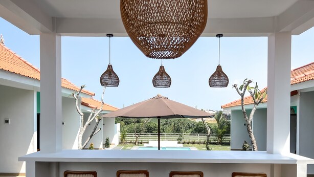 Un área de estar al aire libre con techo y vista a la piscina, con una barra blanca y tres sillas de mimbre.