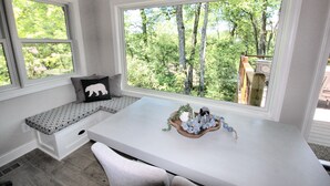 Une pièce avec un bureau blanc, un banc orné d’un coussin en forme d’ours et une grande fenêtre donnant sur une forêt.
