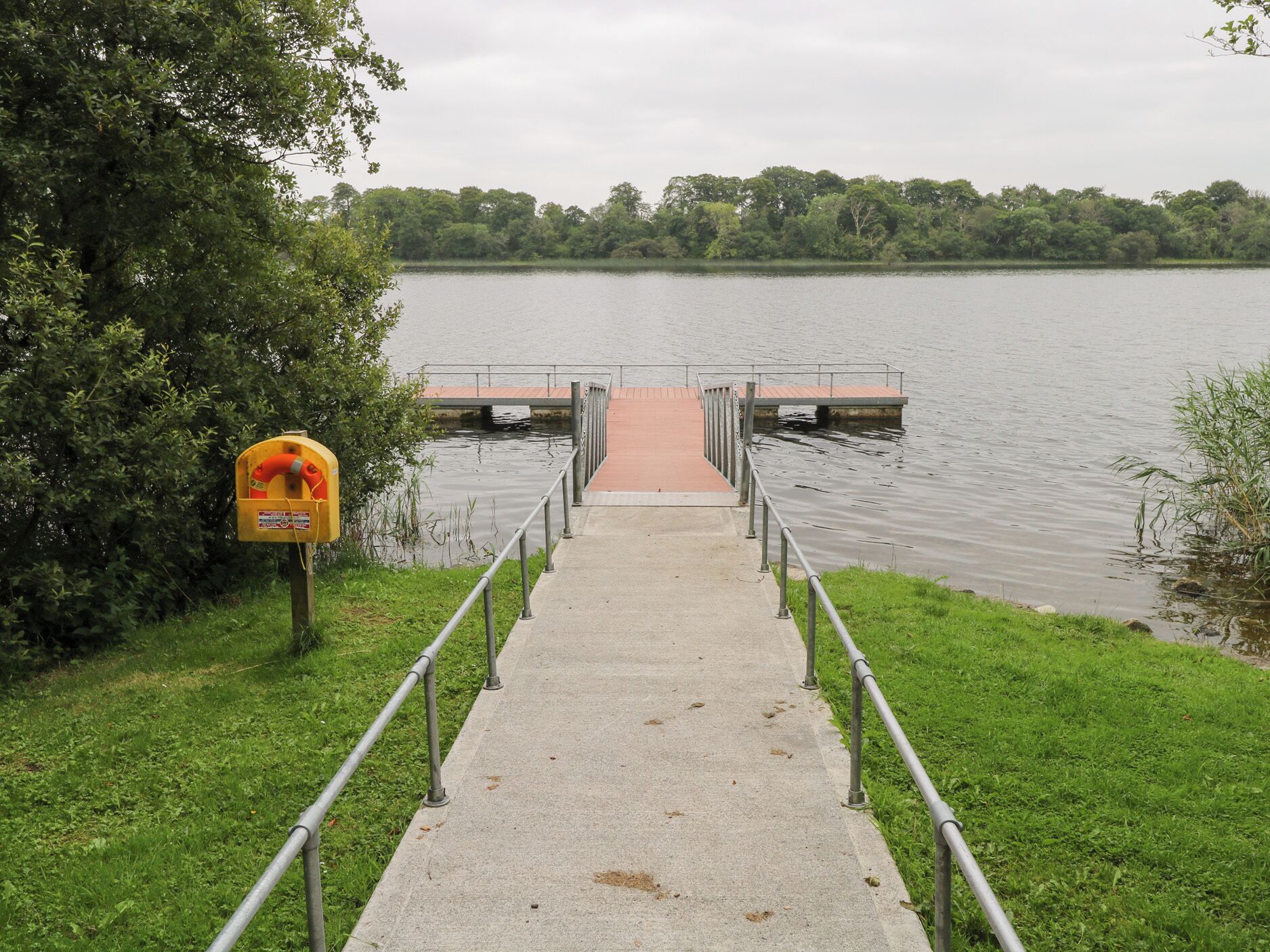 Ein Betonweg führt zu einem Steg über dem Wasser, links ein Rettungsring und rechts eine Rasenfläche.