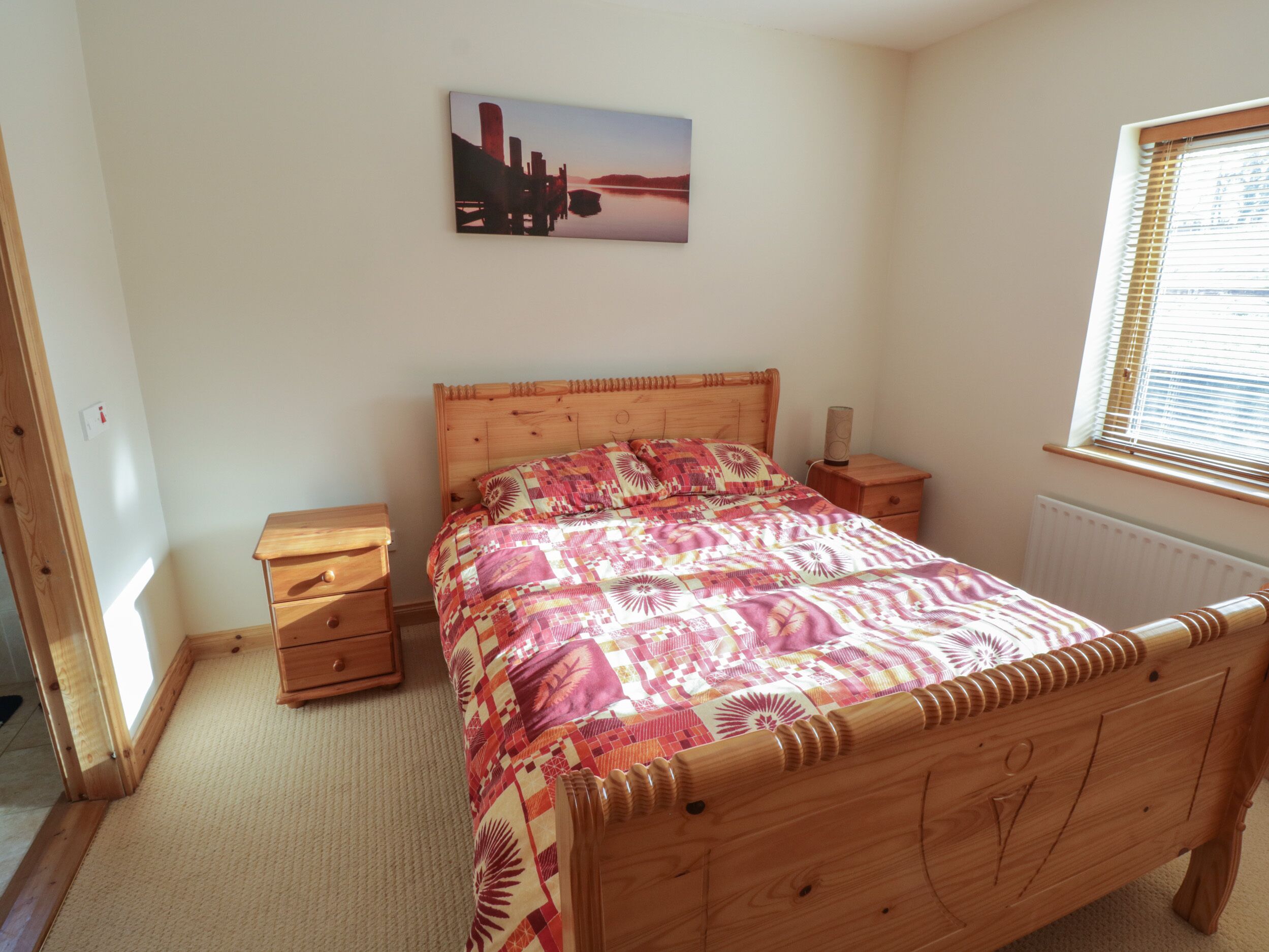 A bedroom with a wooden bed, a bedside table, a nightstand, a radiator, and a window with blinds.