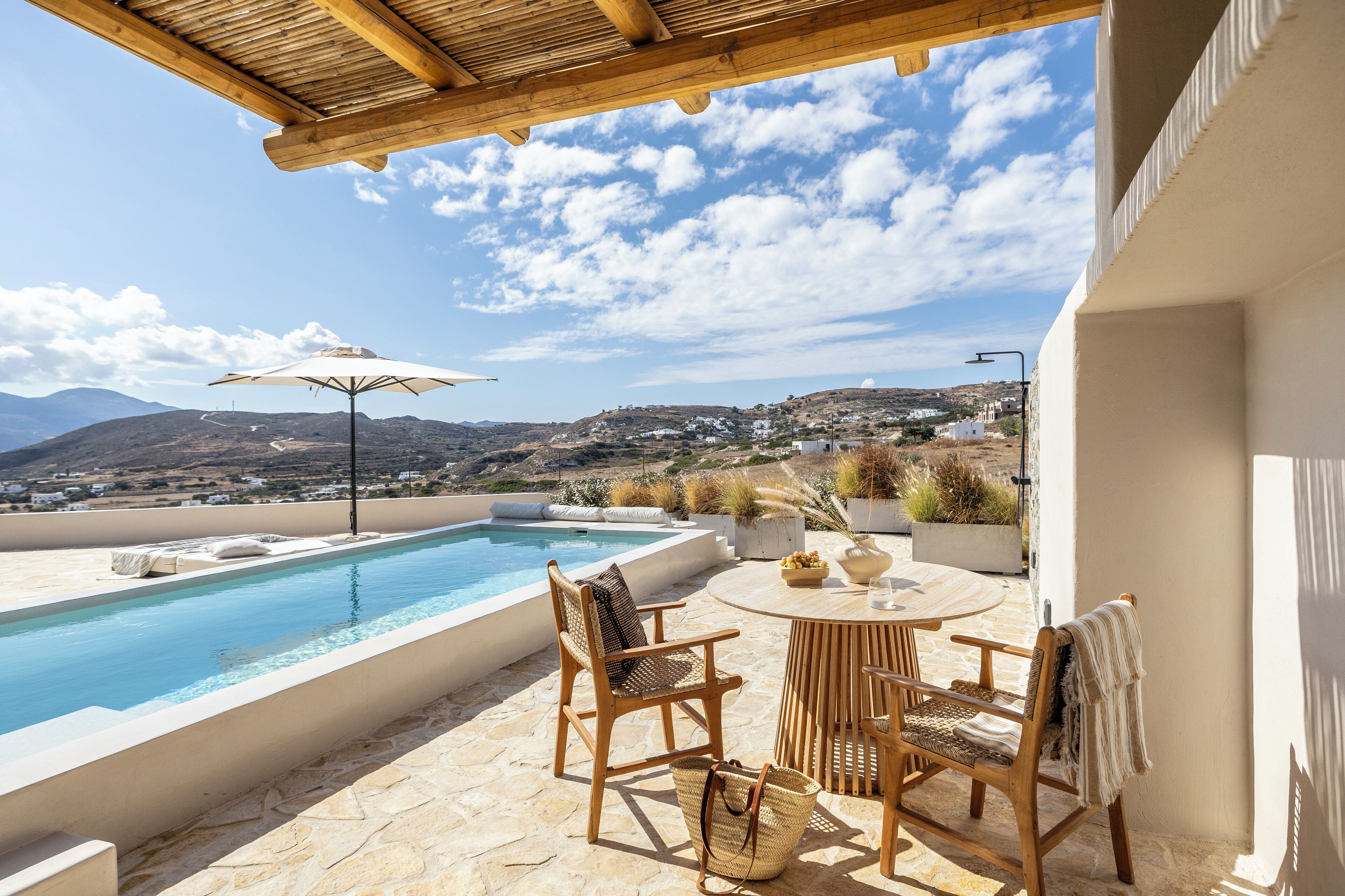 A poolside area with a wooden pergola, two wicker chairs, a table, and a view of a hillside town.