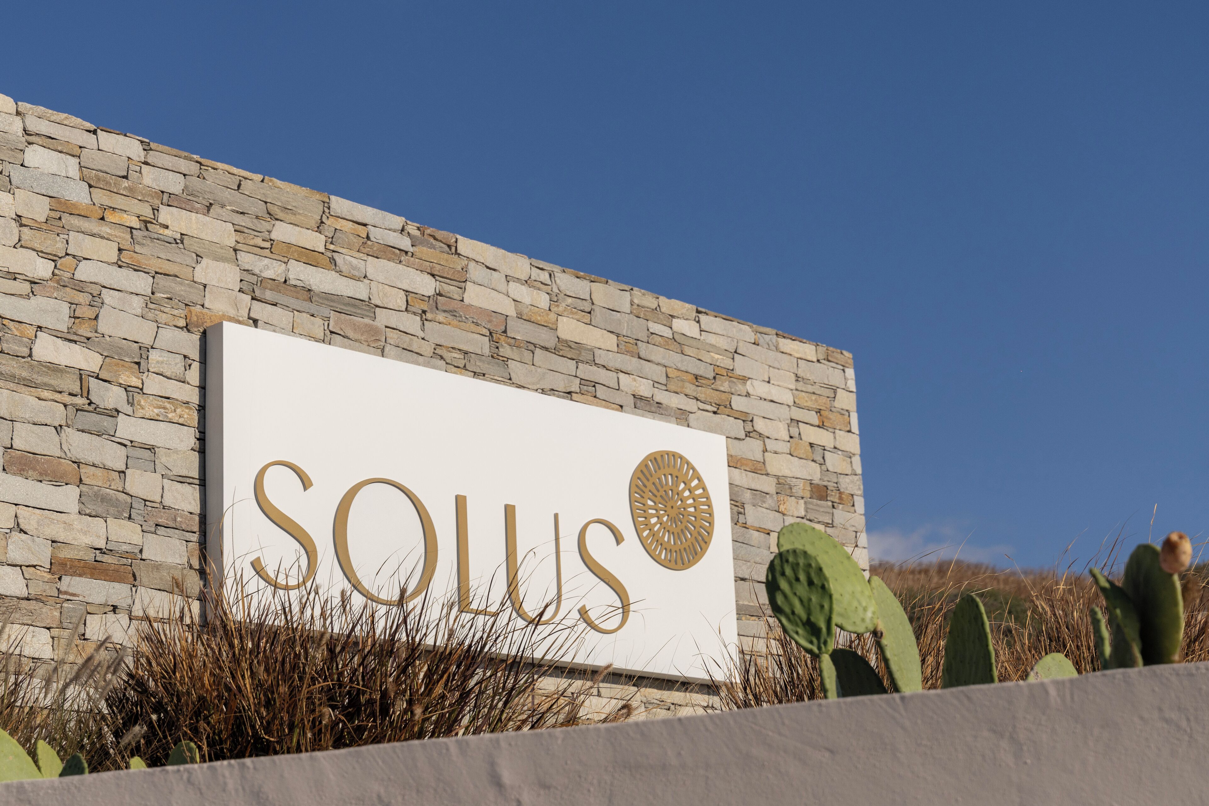 A sign with the word "SOULUS" and a circular design, set against a stone wall with cacti in the foreground.