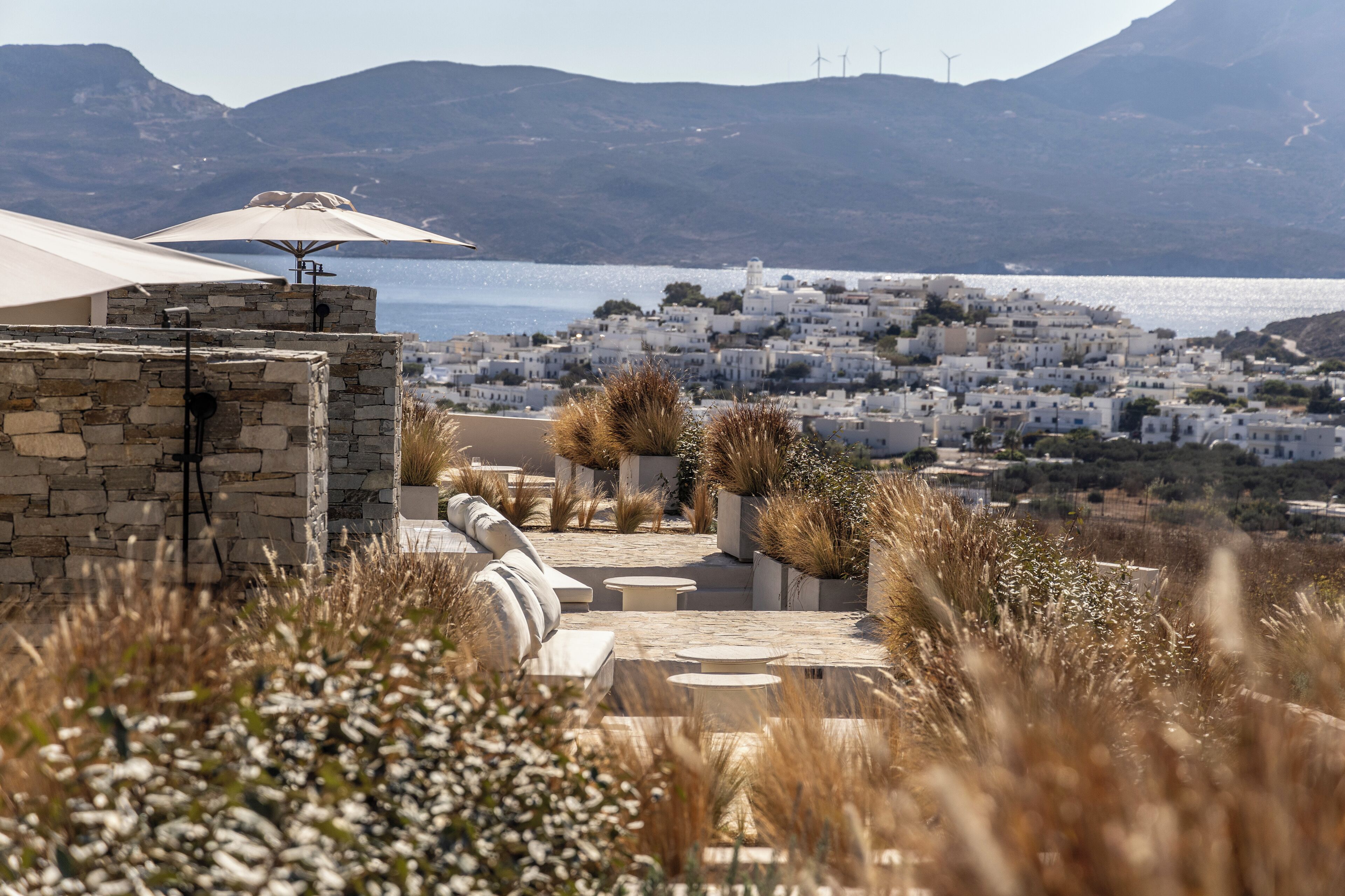 A terrace with stone walls, outdoor seating, and a view of a coastal town with white buildings and wind turbines in the distance.
