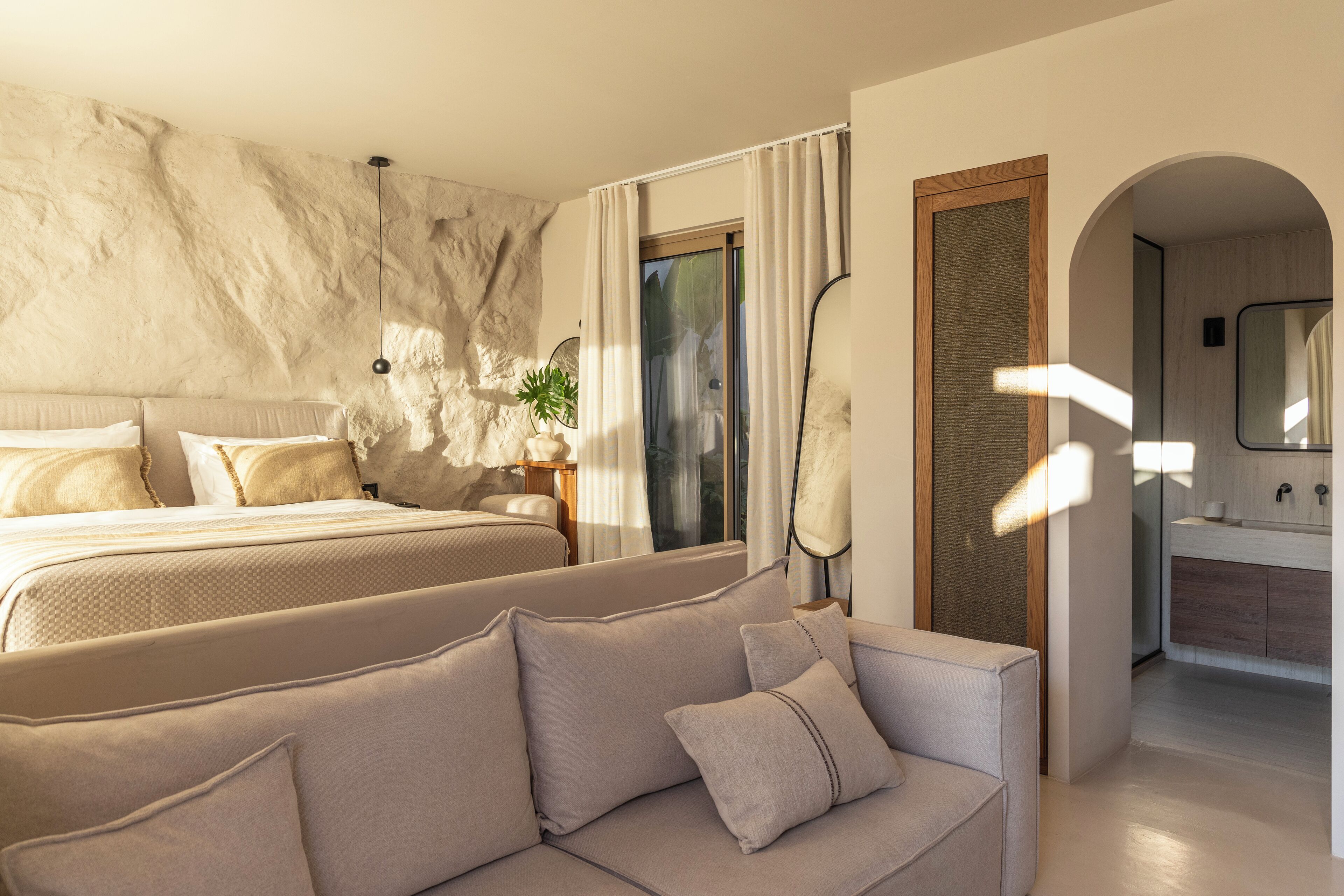 Un dormitorio moderno con una cama grande, un sofá, un espejo y un baño visible a través de un arco.