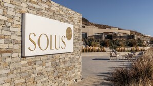 A sign with the word "SOLUS" on a stone wall, a modern building in the background, and outdoor seating area. - Solus Suites (Milos)