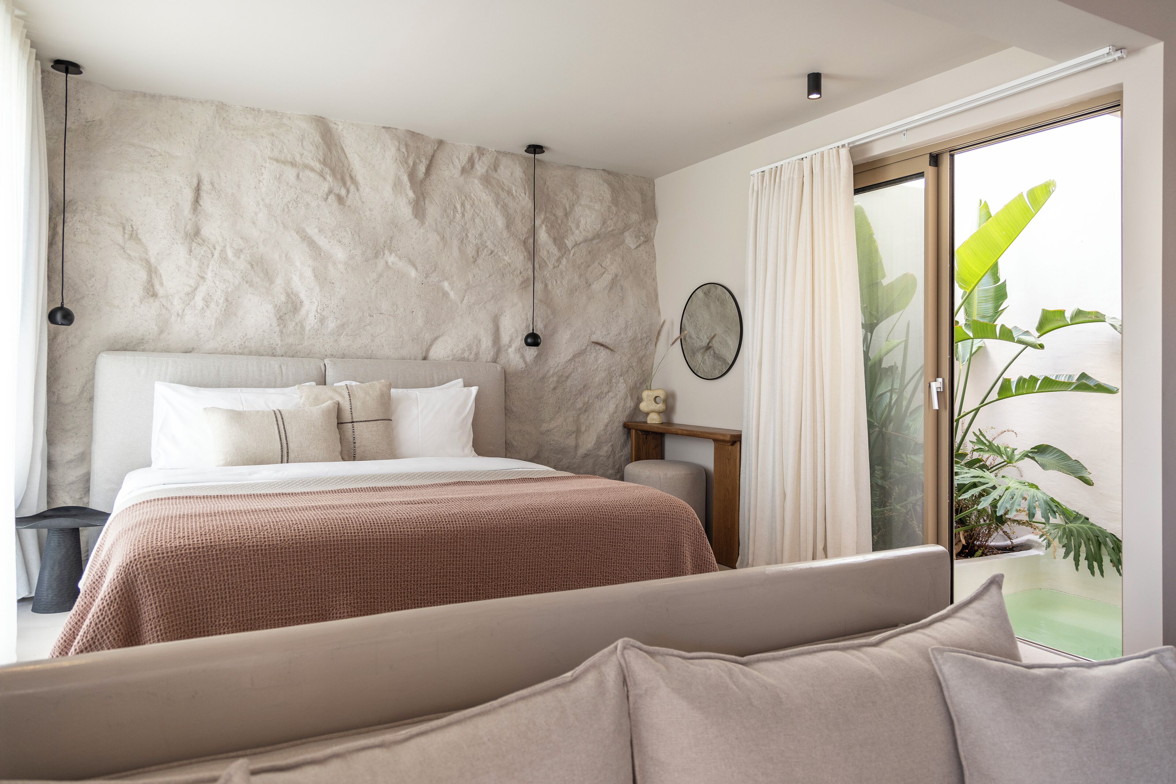 Ein Schlafzimmer mit einem großen Bett, einem Nachttisch, einer Wanduhr und einer Glastür, die auf einen Balkon mit Pflanzen führt.