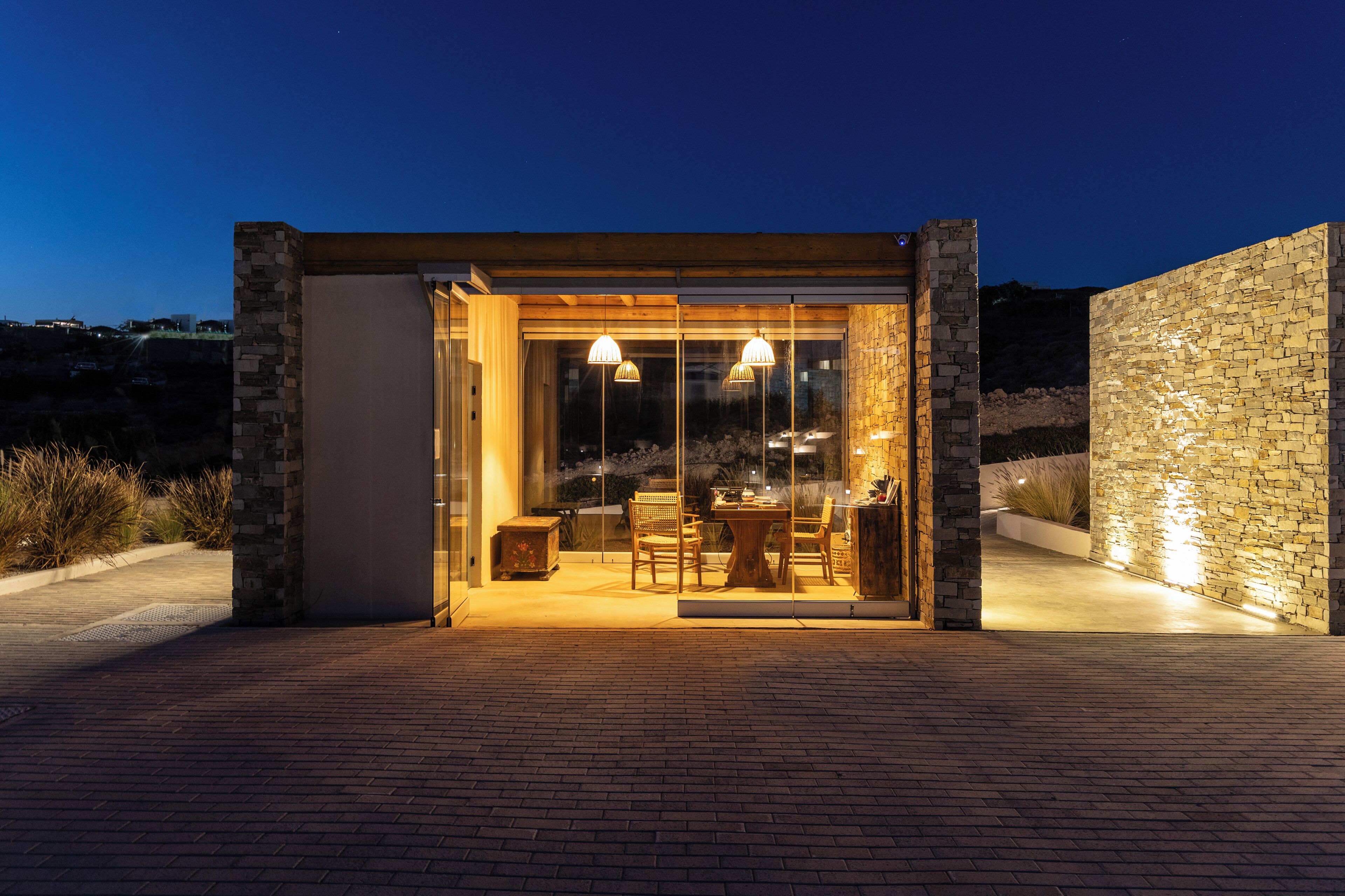 A modern outdoor dining area with a glass enclosure, stone walls, and ambient lighting.