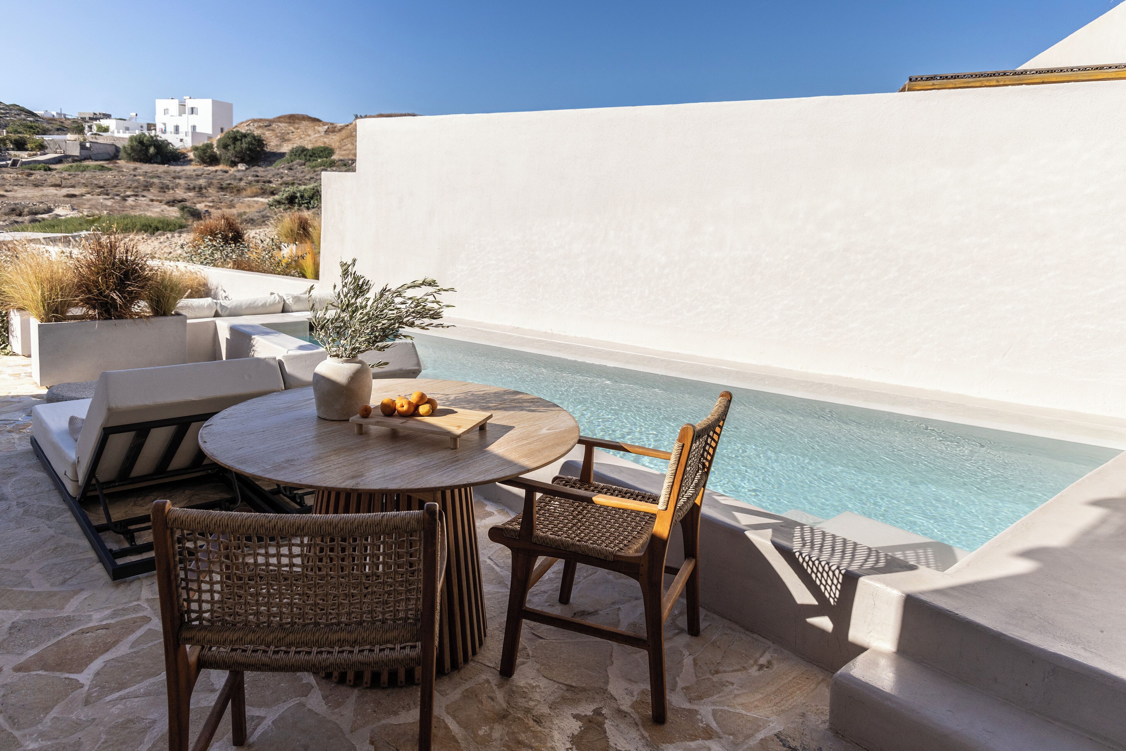A poolside area with a round table and wicker chairs, overlooking a pool and a white building in the background.