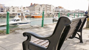 A bench by a canal with boats and buildings in the background. - Mellstock (Weymouth)