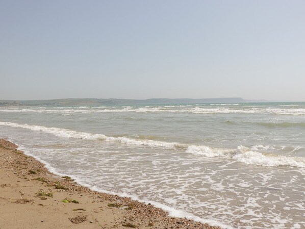 A beach with waves gently lapping at the shore, a clear sky, and distant hills. - Mellstock (Weymouth)