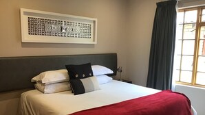 Una cama bien hecha con ropa blanca y una manta roja, un cabecero, una obra enmarcada sobre la cama y una ventana con cortinas.