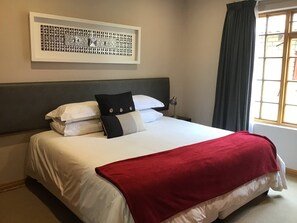 Cama bem arrumada com roupa de cama branca e manta vermelha, cabeceira, quadro na parede acima da cama, e janela com cortinas.