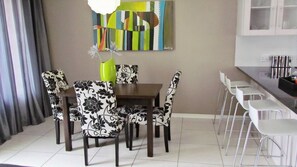 A dining area with a table and chairs, a kitchen area with a counter and chairs, and a wall with a colorful abstract painting. - Comfortable Furnished Apartment Wi-fi Free Parking (Bloemfontein)