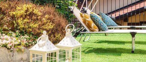 Uma espreguiçadeira com almofadas estampadas, um candeeiro branco e um jardim com relva verde e arbustos.