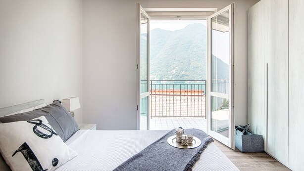 Un dormitorio con cama, mesita de noche, ventana con vistas a las montañas y balcón con barandilla.