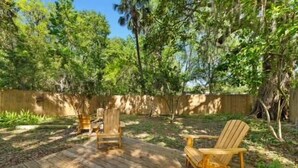 A wooden deck with two Adirondack chairs and a small table, surrounded by lush greenery and trees. - Cozy Cottage | Fenced Yard & Fire Pit (Homosassa)