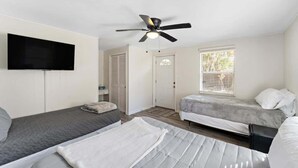 A compact room with two beds, a ceiling fan, a TV mounted on the wall, and a window with blinds. - Cozy Cottage | Fenced Yard & Fire Pit (Homosassa)