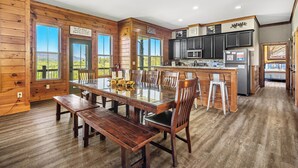 A spacious dining area with a large wooden table and benches, a kitchen with dark cabinets, and large windows offering a view of the outdoors.