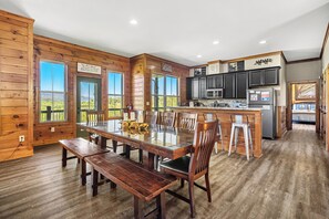 Enjoy meals at the large dining table surrounded by comfortable chairs, with a kitchen area and large windows offering a view of the outdoors.
