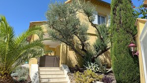 A yellow building with a staircase leading to a door, surrounded by various plants and a tall tree. - Villa Ocre, in Provence Verte, between the Mediterranean and the Gorges du Verdon. (Lorgues)