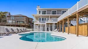 Zona de piscina con terraza de madera, sillas de descanso y un edificio de dos pisos con balcones.