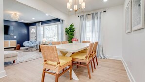 A dining area with a white table and wicker chairs, a chandelier, and a large window with curtains. - Modern 4BR Home Near MTSU and Restaurants (Murfreesboro)