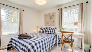 A bedroom with a checkered bedspread, a bedside table with lamps, a chair, and a window with curtains. - Modern 4BR Home Near MTSU and Restaurants (Murfreesboro)