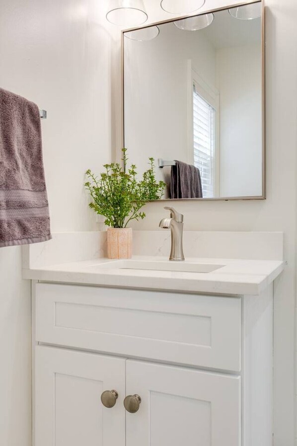 Un baño con tocador blanco, espejo, lavamanos y una planta en maceta.