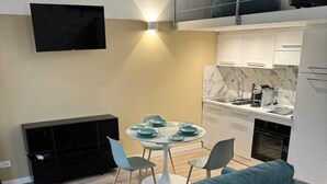 A modern living room with a sofa, a dining table with chairs, a kitchen area, and a TV mounted on the wall.