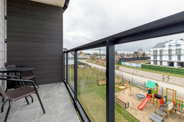 A balcony with a view of a playground, a table with chairs, and a modern building in the background.