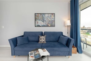 A modern living room with a blue sofa, a framed abstract painting, a lamp, and a view through a window.