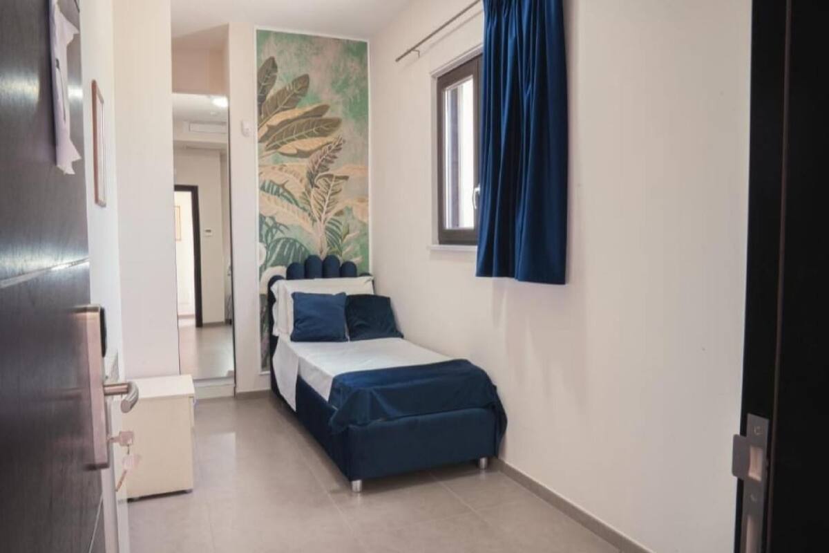 A small, compact room with a bed, a window with curtains, a door, and a wall with a mural.
