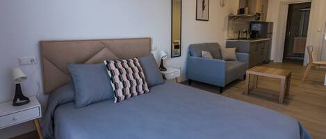 A modern bedroom with a large bed, a bedside table with a lamp, a chair, a small table, and a kitchenette in the background.