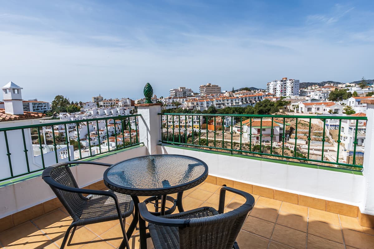 A balcony with a round table and two chairs, overlooking a coastal town with white buildings and a clear sky.