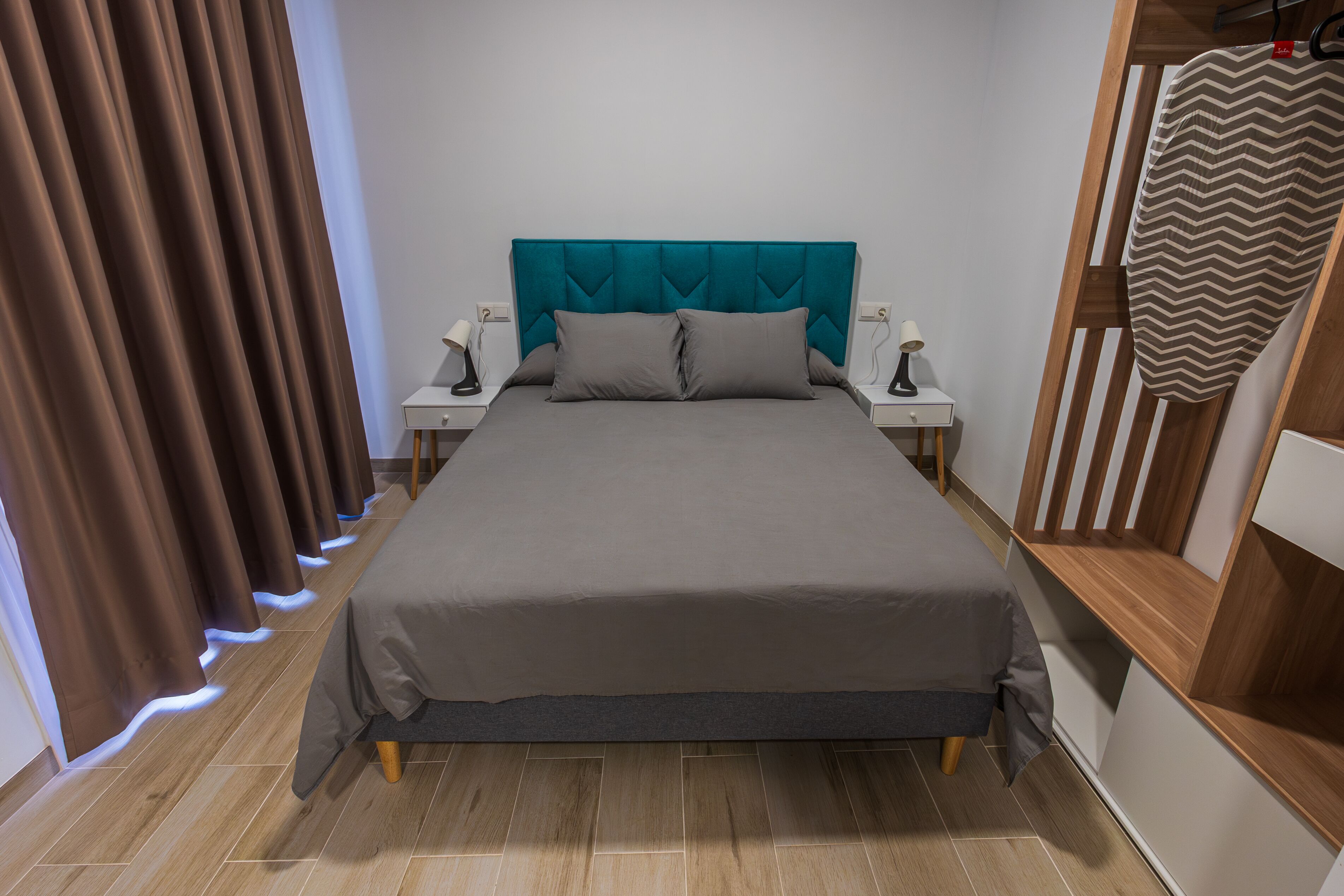 A neatly made bed with a teal headboard, two bedside tables with lamps, and a wooden shelf unit.