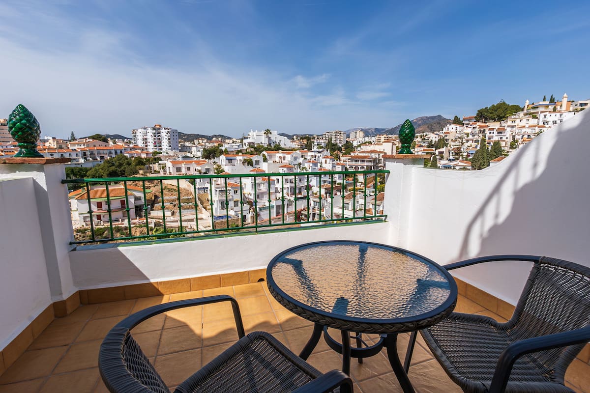 A balcony with a round table and two chairs, overlooking a residential area with white buildings and green trees.