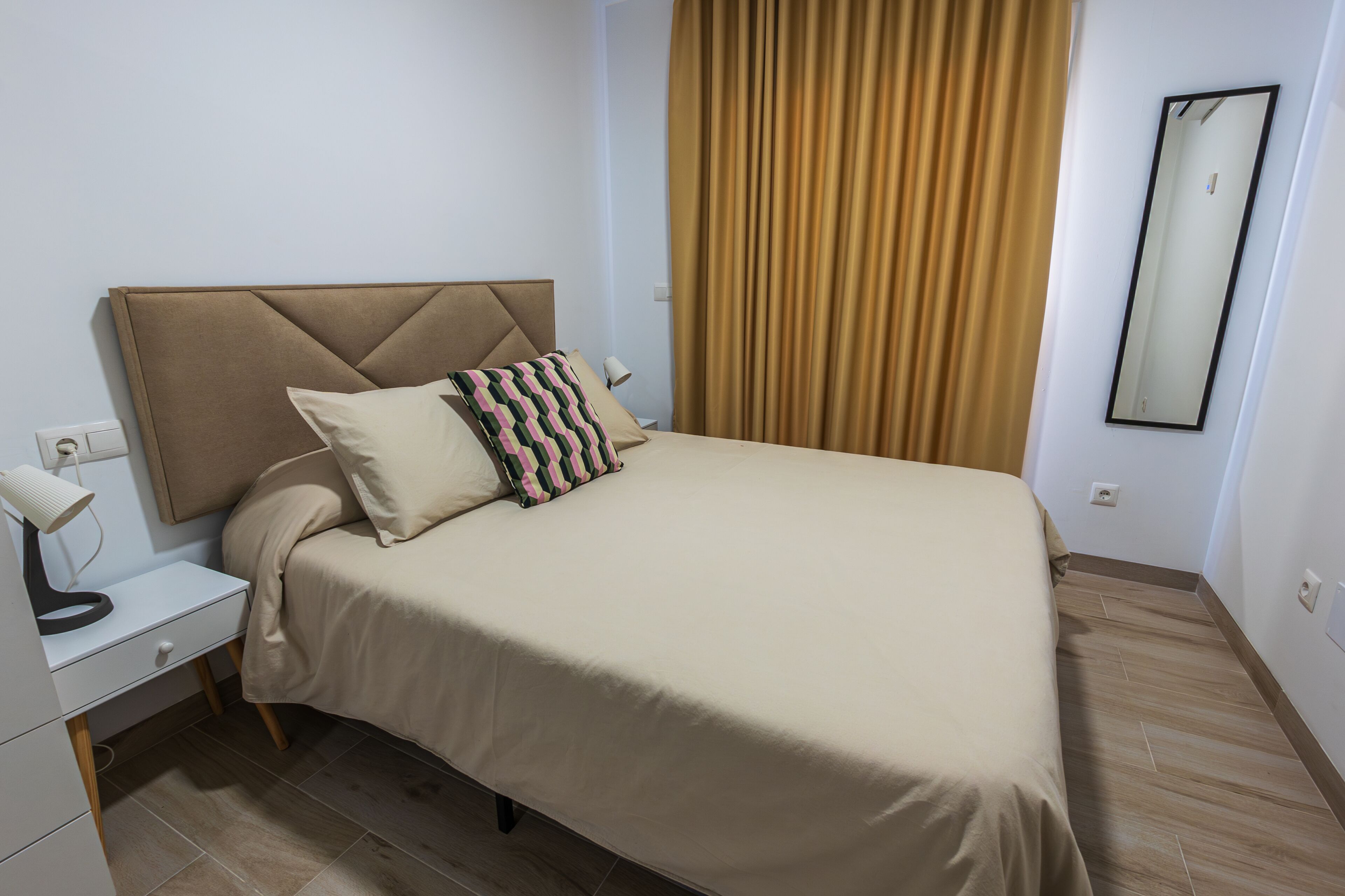 Een netjes opgemaakt bed met hoofdbord, nachtkastje met lamp en spiegel aan de muur.