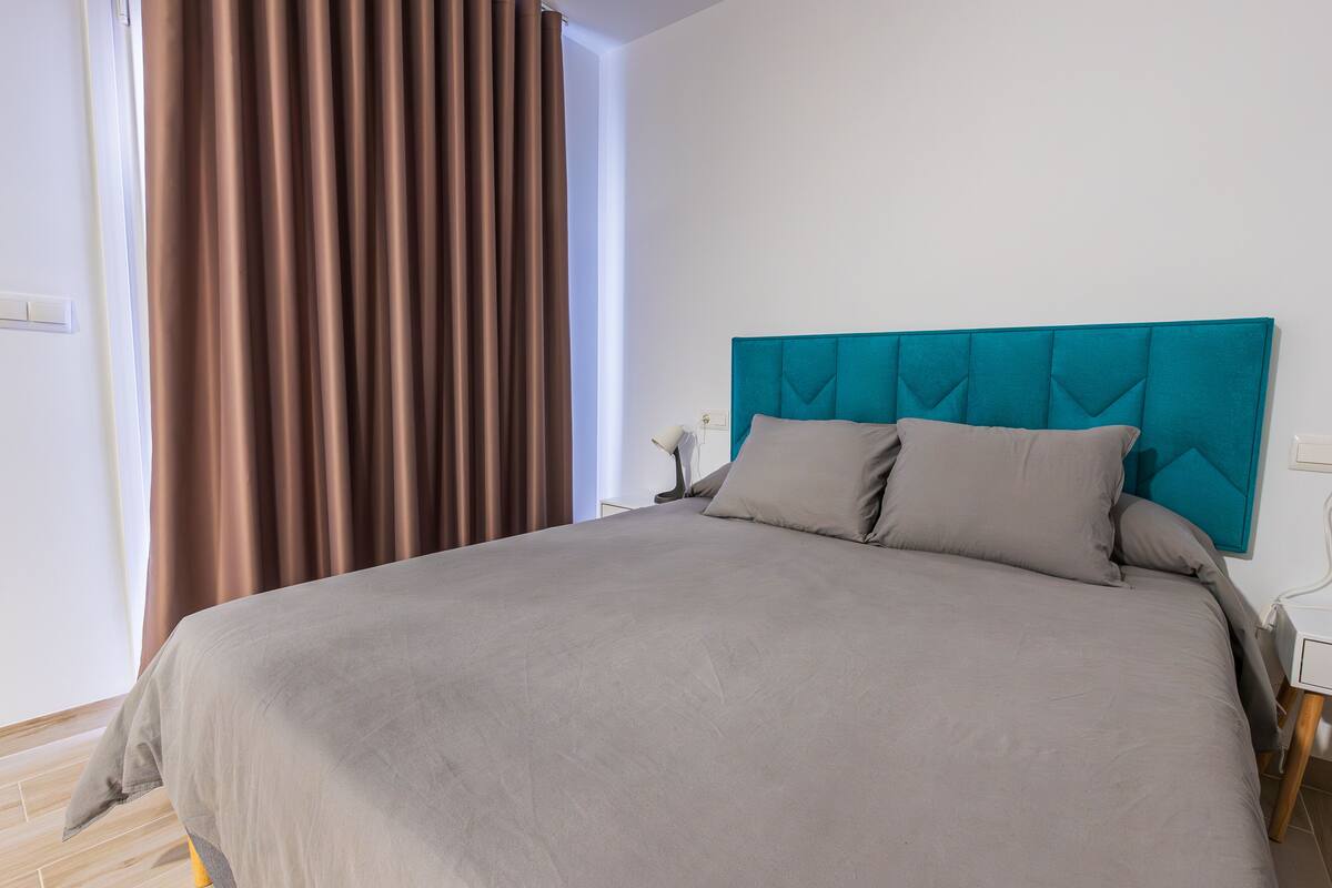 A modern bedroom with a large bed, a teal headboard, and a view of the outdoors through a window.