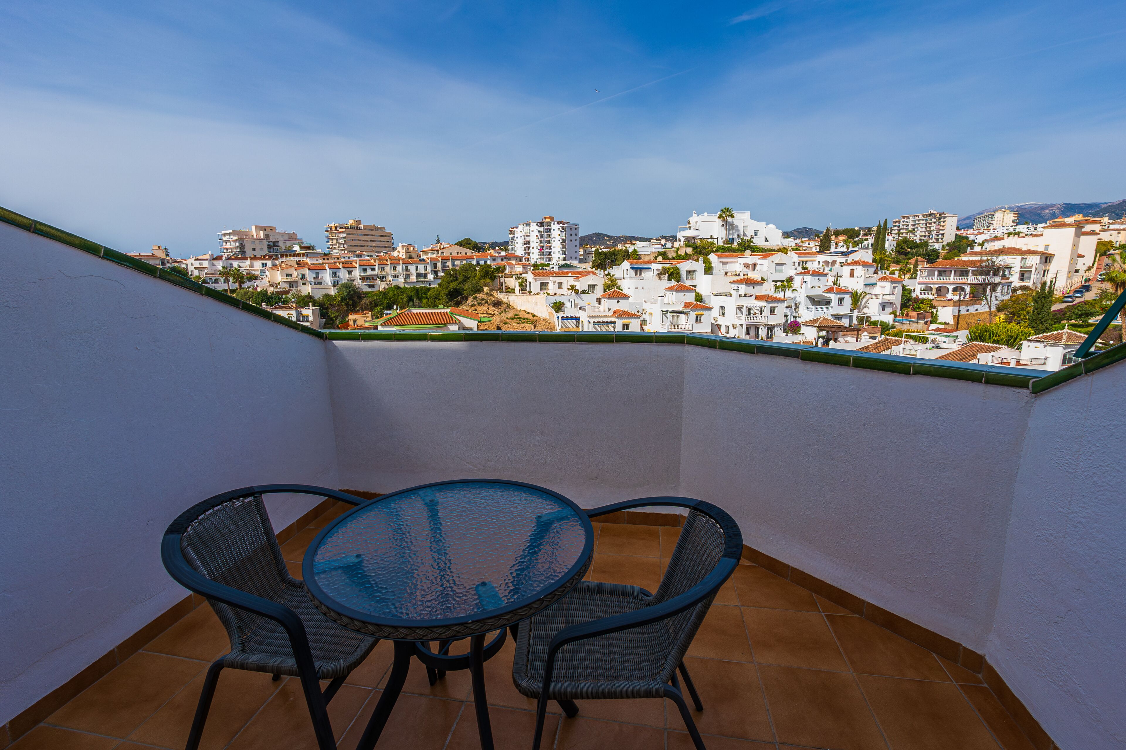 A balcony with a table and chairs, overlooking a residential area with white buildings and red roofs.