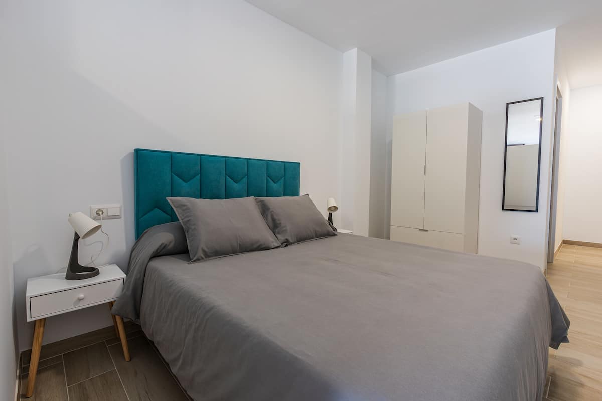 A neatly made bed with a teal headboard, a bedside table with a lamp, and a wardrobe in a modern, minimalistic room.