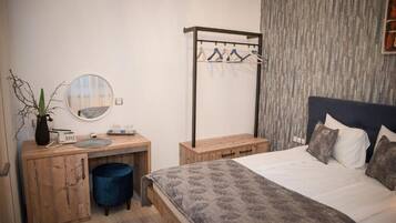 A bedroom with a bed, a wooden desk with a mirror, a hanging clothes rack, and a blue waste bin.