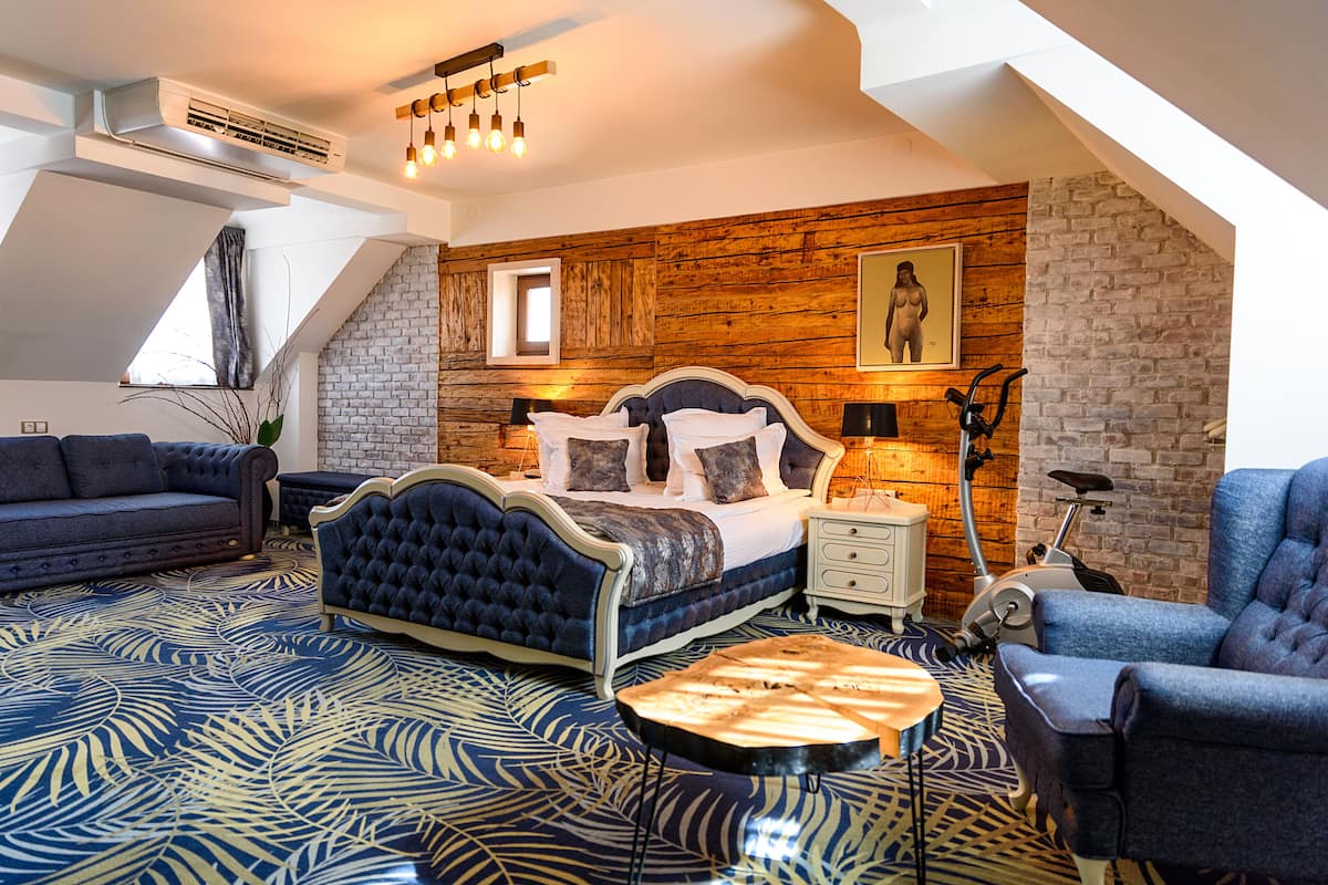 A bedroom with a loft-style ceiling, a bed with a headboard, a nightstand, a lamp, a chair, and a floor with a leaf-patterned carpet.