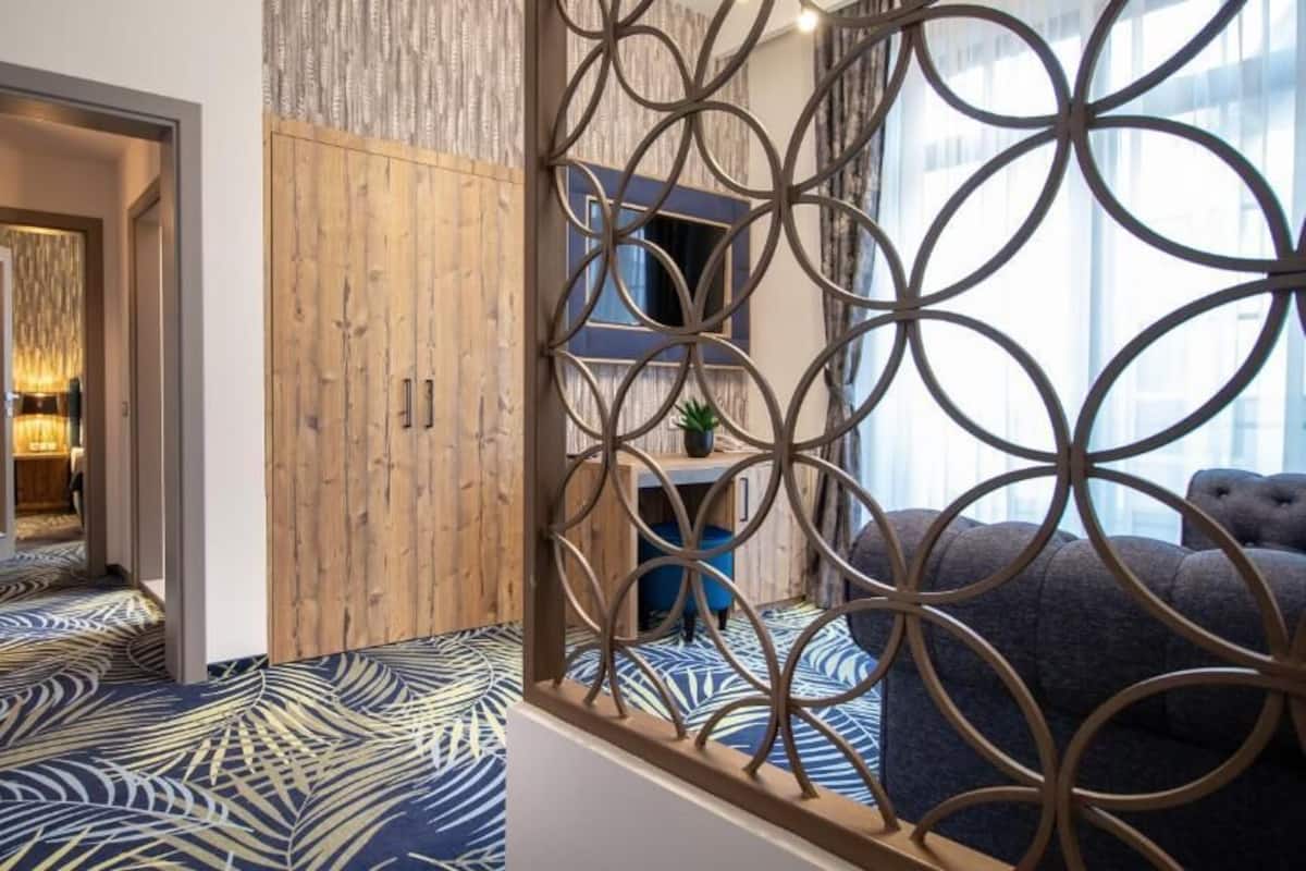 A hotel room with a patterned carpet, a wooden wardrobe, a decorative metal partition, and a view of the city through large windows.