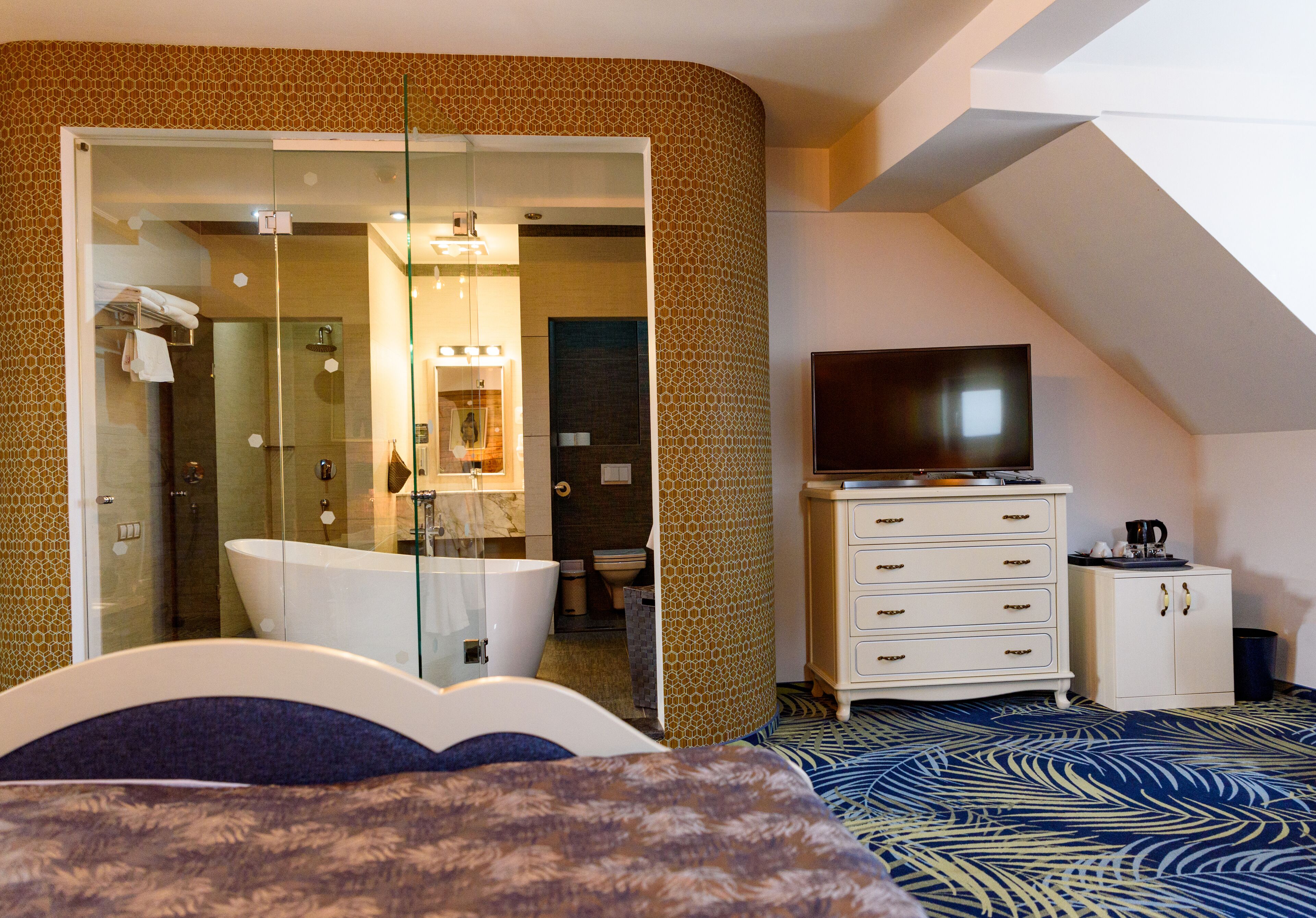 A modern hotel room with a glass-enclosed shower, a flat-screen TV, a white dresser, and a patterned carpet.
