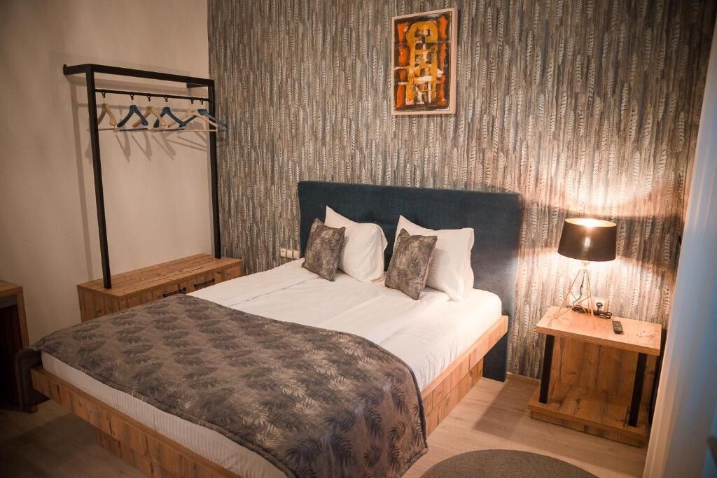 A bedroom with a bed, a wooden nightstand, a wall-mounted lamp, and a clothing rack.