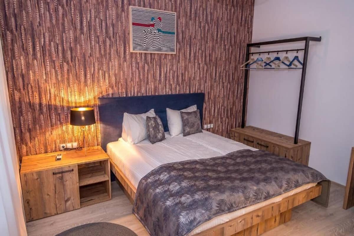 A bedroom with a wooden headboard, a bed with a patterned blanket, a bedside table with a lamp, and a wall-mounted coat rack.