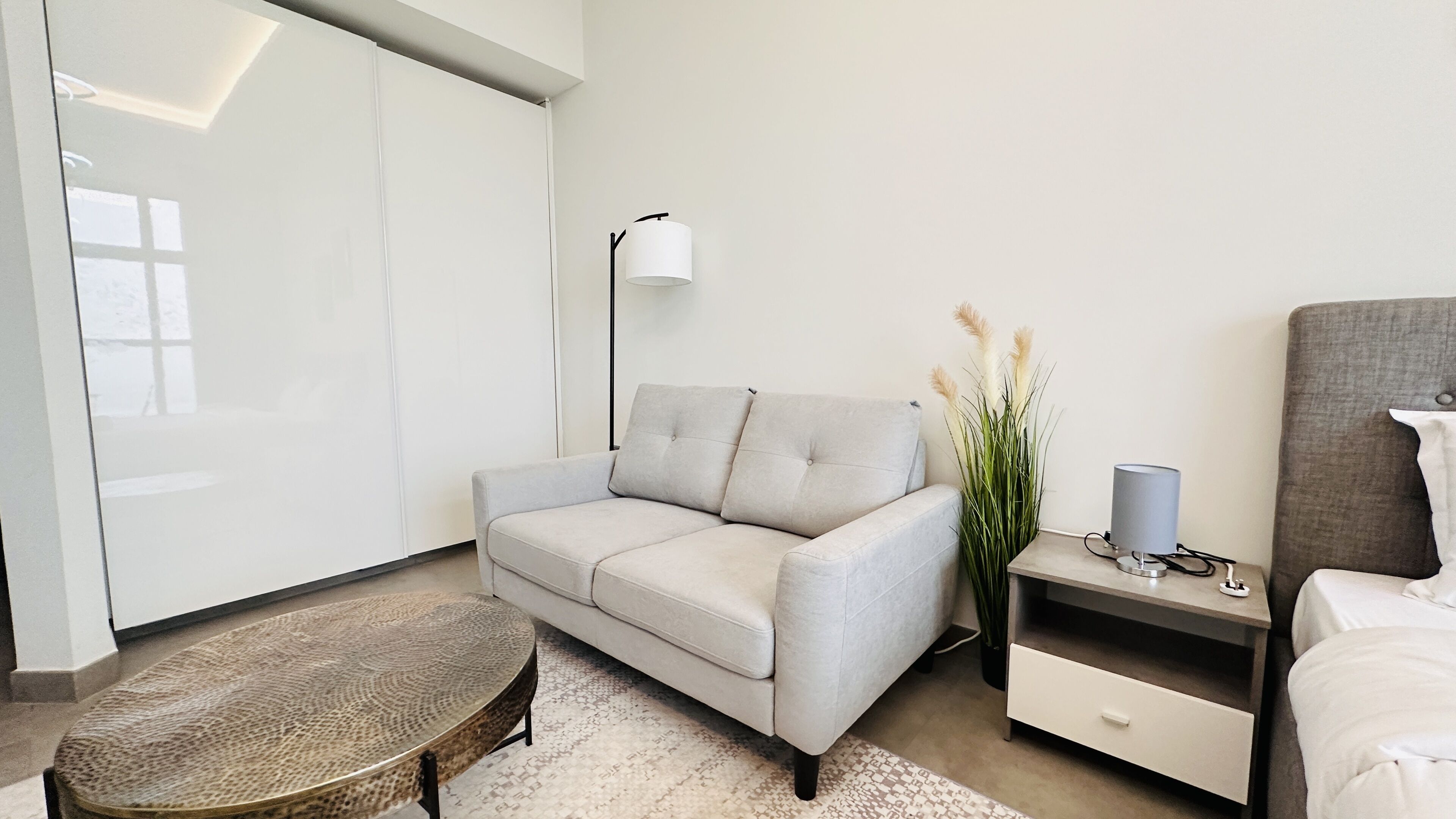 A modern hotel room with a grey sofa, a small round table, a bedside table with a lamp, and a bed.