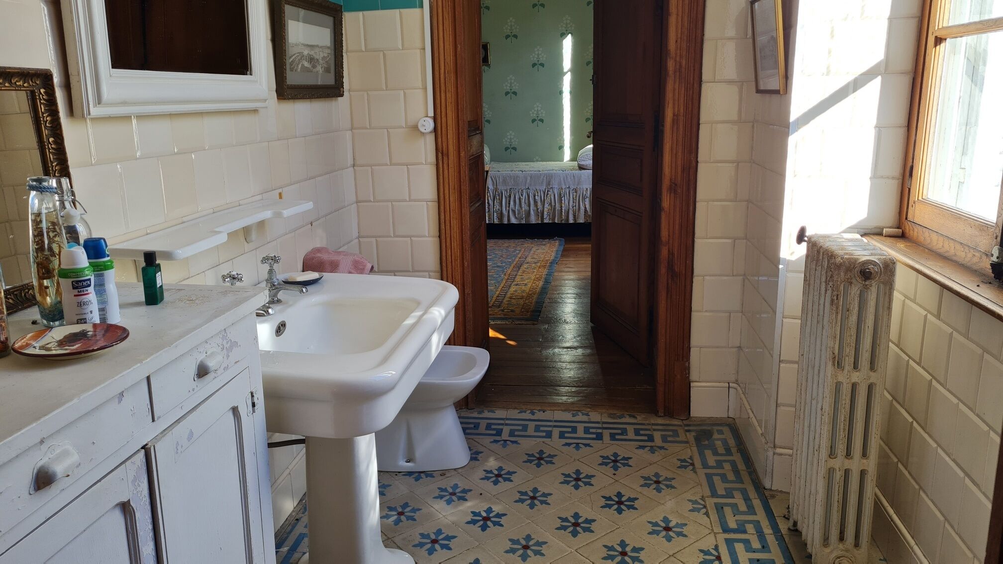 Un baño estrecho con lavamanos, inodoro y espejo. Hay una ventana a la derecha y una puerta que da a otra habitación.