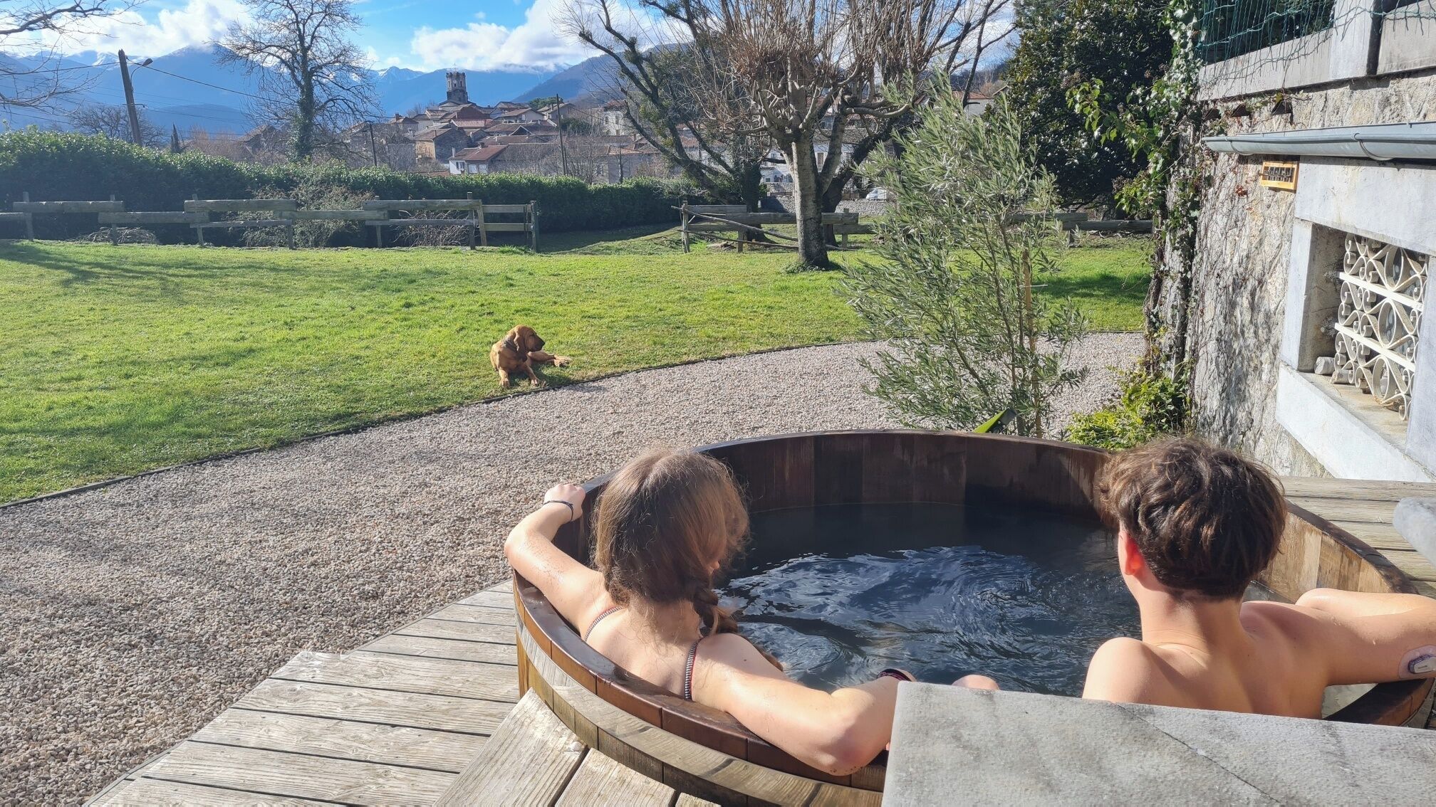 Una vasca idromassaggio in legno con due persone sedute all'interno, affacciata su una zona erbosa con un cane e un edificio in pietra sullo sfondo.