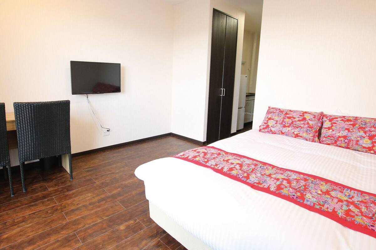 A hotel room with a bed, a TV mounted on the wall, two chairs, and a wardrobe.